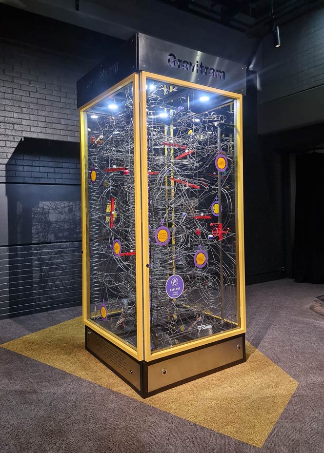 Kinetic sculptures lift, roll and drop billiard balls around various pathways. A large green steel and glass display case sits within a white foyer. Within the display case is a series of wire pathways and billiard balls.
