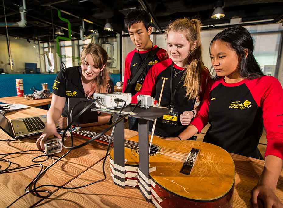 Four people looking at a robotic guitar