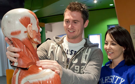 A man a woman smiling, wearing track suit tops are looking and handling a model human skeleton with muscles on it.