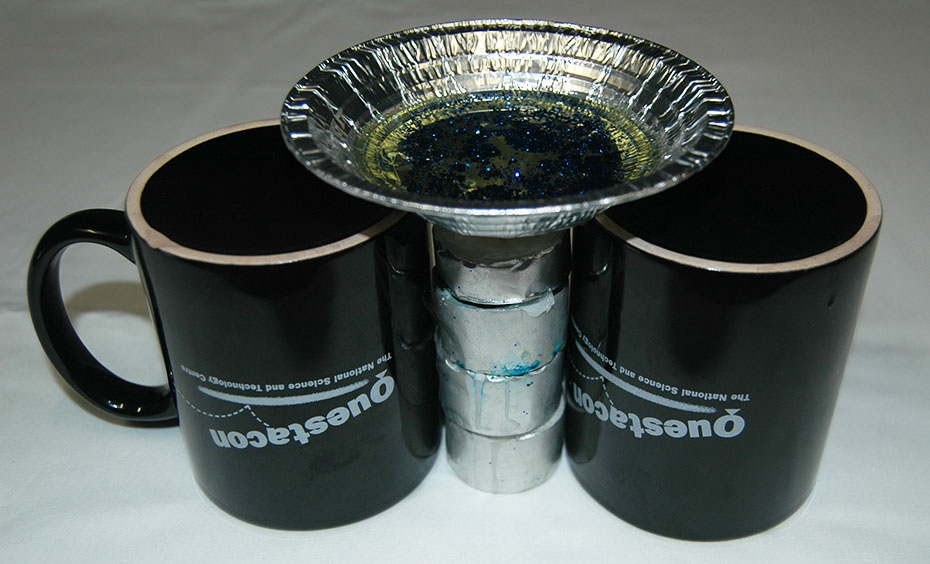 This experiment requires adult supervision. Perform this experiment away from curtains or other flammable materials. two black upside down mugs on a white table cloth, with a silver cylinder between them, and a small aluminium dish sitting on the cylinder