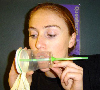 Play your handmade honker by blowing through the hole in the side of the cup. A woman with her mouth resting on a clear plastic cup. The cup as a green straw running through the length of it, with a latex glove over the cup mouth.