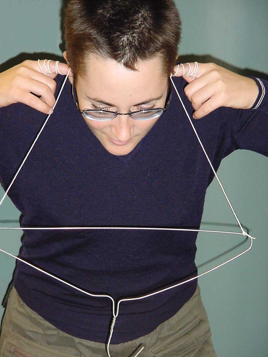 With the string still wrapped around your fingers, place your index fingers in your ears and bend over so the coat-hanger doesn’t touch anything. A woman wearing glasses looking down, with her index fingers held to her ears, and white string rapped around her fingers drops down to hold up an upside down wire coat hanger.
