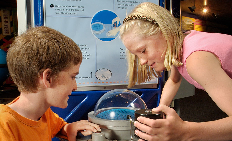 Use the button and hand pump to remove air from the glass dome and watch how the rubber sheet responds. A boy and girl operating a button and watching a clear dome with weights attached. In the background is a blue and white information panel.