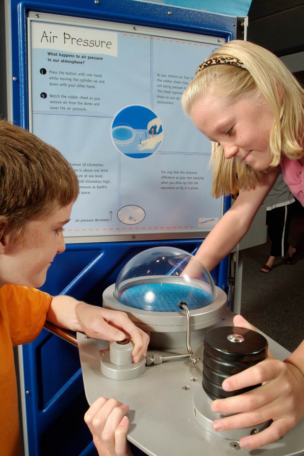 Air pressure changes as you travel higher into Earth's atmosphere. A boy and girl operating a button and watching a clear dome with weights attached. In the background is a blue and white information panel.