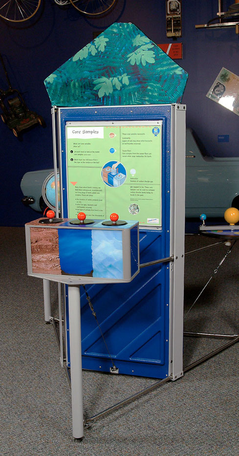 Scientists take core samples to study layers of ice or earth and their age, composition and evidence of living organisms. A blue, green and grey exhibt which has 3 core sample boxes at the front with red knobs on top of each, and a large vertical information panel at the back.