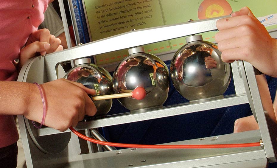 Tap each globe and listen carefully. Look at the base of each globe to check what is inside. A boy and girl standing in front of a green and blue vertical information panel have their hands on a grey framed exhibit, which has 3 chrome spheres. The girl has a small mallet, and is hitting the centre sphere.