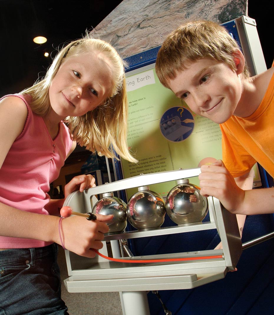 Scientists can explore what is inside the Earth by studying vibrations. A boy and girl standing in front of a green and blue vertical information panel have their hands on a grey framed exhibit, which has 3 chrome spheres. The girl has a small mallet.