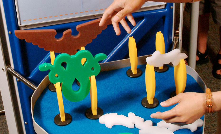 Insert the eight corn pieces into the slots and balance the mice and snake pieces on top. A blue and grey exhibit table and matching information panel with red, green, white and yellow plastic animal shapes on the table top.