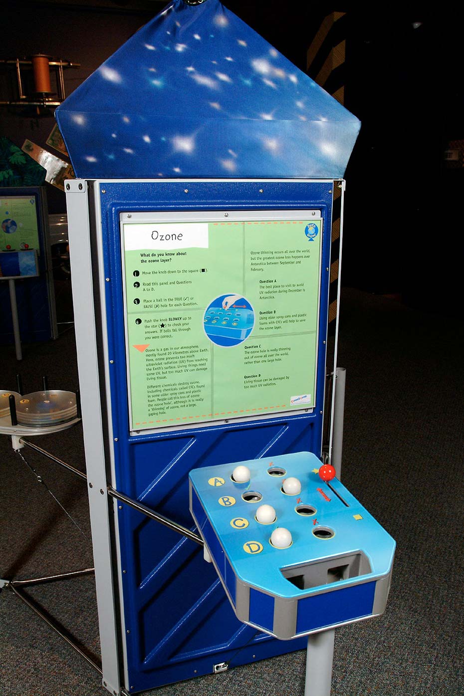Ozone is a gas in our atmosphere that prevents too much ultraviolet radiation (UV) from reaching the Earth's surface. A blue and green exhibiti with a vertical information panel at the back and a horizontal table with white balls, a red lever and the letters 'A', 'B', 'C' and 'D' displayed.