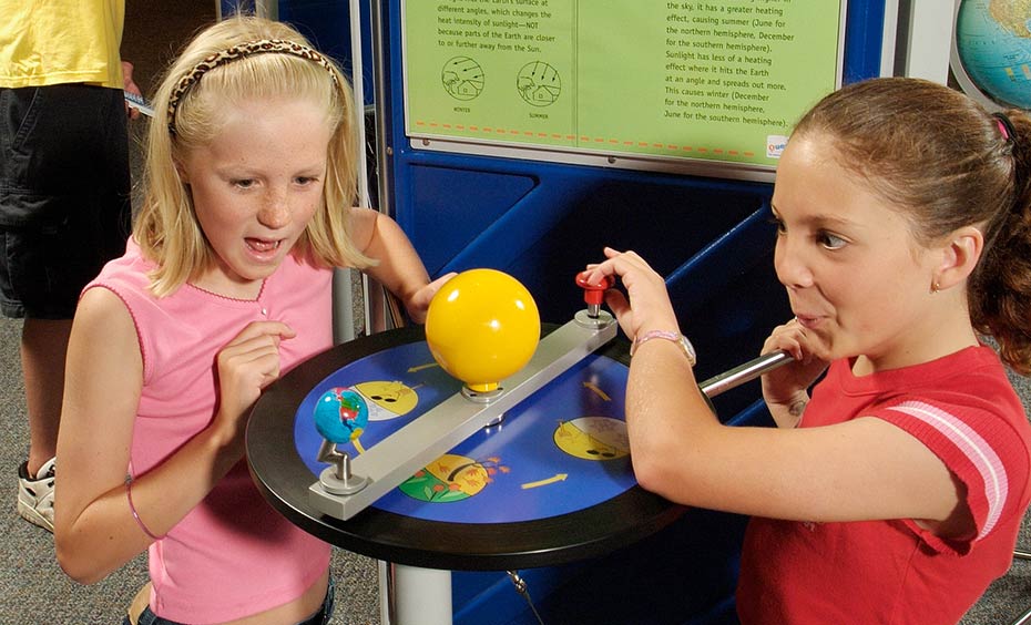 Move a model of planet Earth (tilted on its axis) in orbit around the Sun and notice how Earth's tilt would create different angles of incidence as sunlight hits the ground. Two girls kneeling in front of a blue and green table and information panel. On the table is a yellow model sun and blue earth.