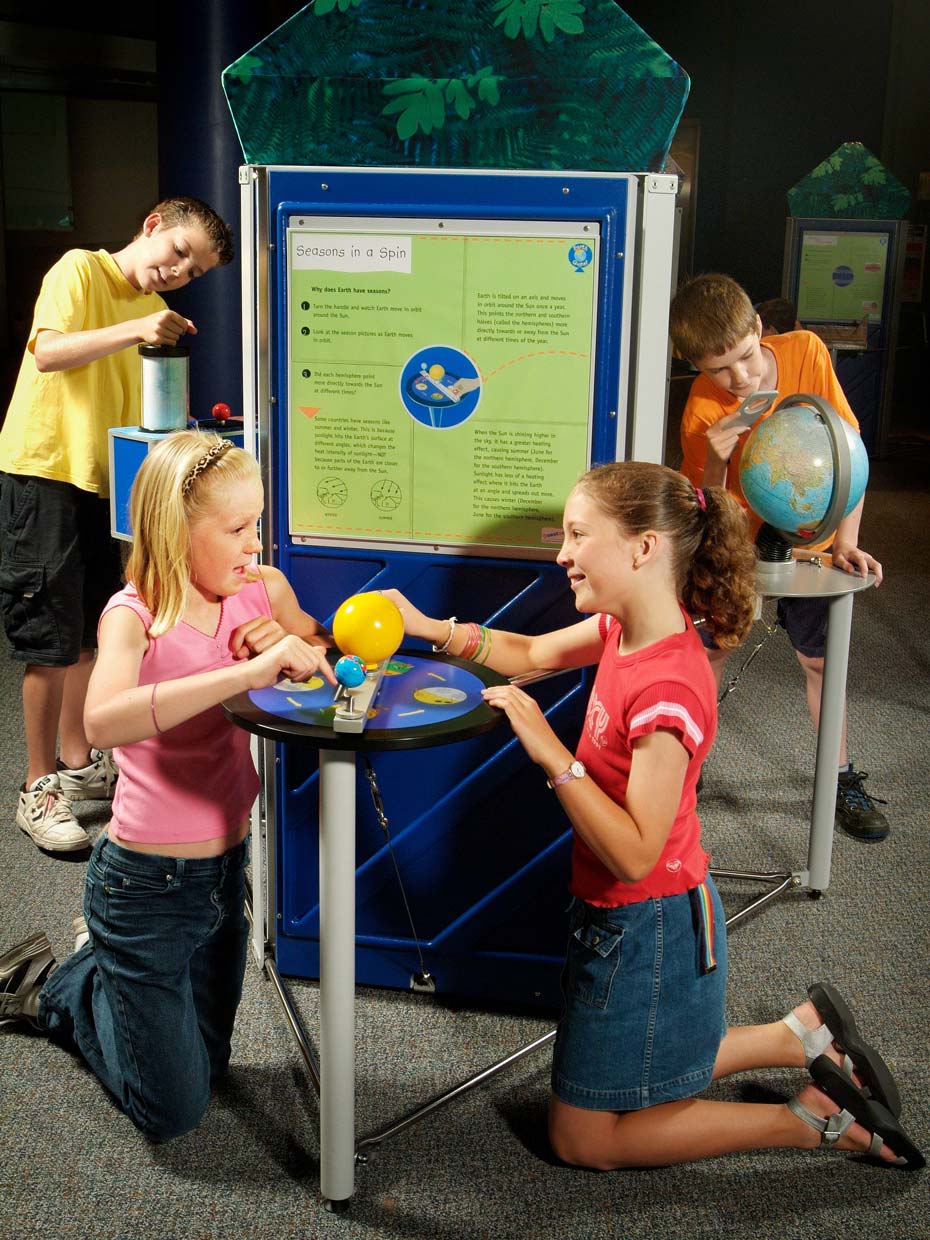 Seasons are caused by the different angles at which sunlight hits the Earth's surface. Two girls kneeling in front of a blue and green table and information panel. On the table is a yellow model sun and blue earth. In the background are two boys looking at other nearby exhibits.
