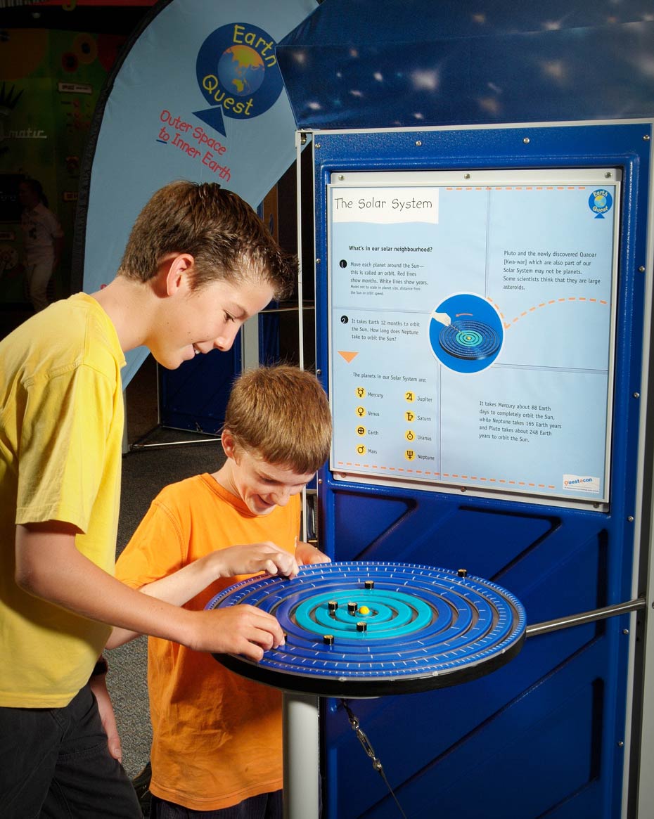 Planets in the Solar System orbit around the Sun, with planets furthest away from the Sun taking many years to complete one orbit. Two boys are playing with a blue disc with black markers that represents the solar system. In the background is a blue and white information panel.