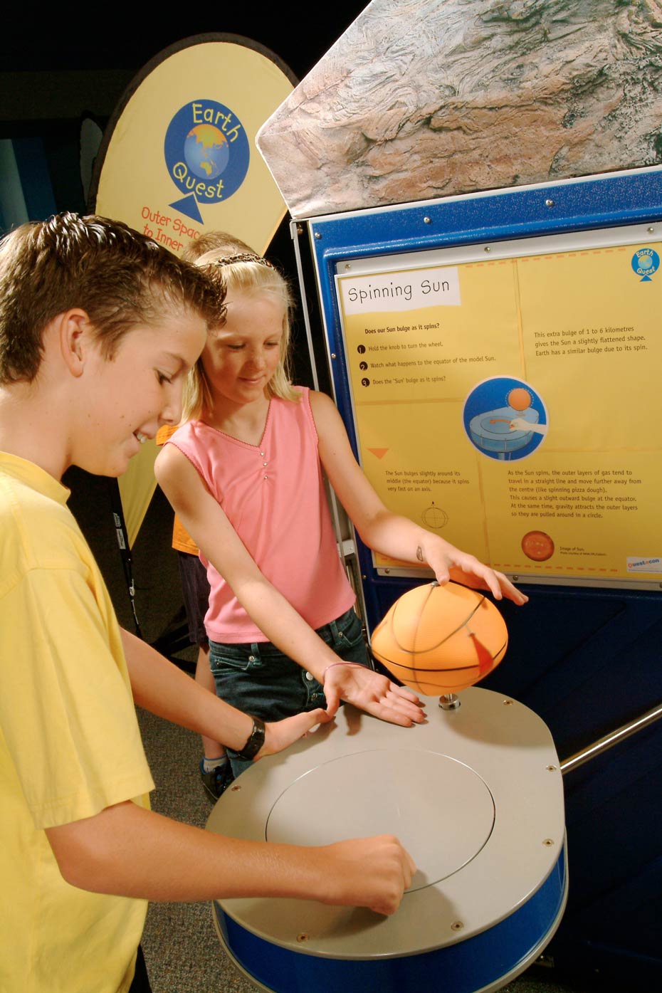 The Sun spins, causing it to bulge around its equator. A boy and girl standing next to a blue and beige round table that has a small basket ball on top. In the background are yellow and blue information panels.