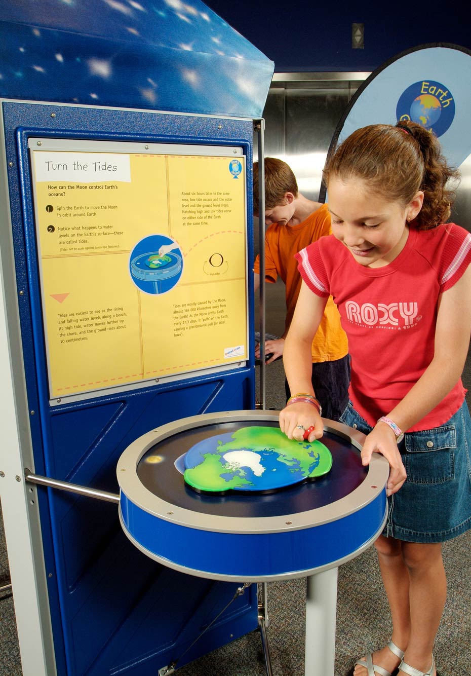 Tides are mostly caused by the Moon and solid earth also responds to tidal movements. A young girl is standing next to a yellow and blue table and exhibition panel. She is winding a red handle on the surface of the table which in turn is attached to a flat blue, white and green earth picture.
