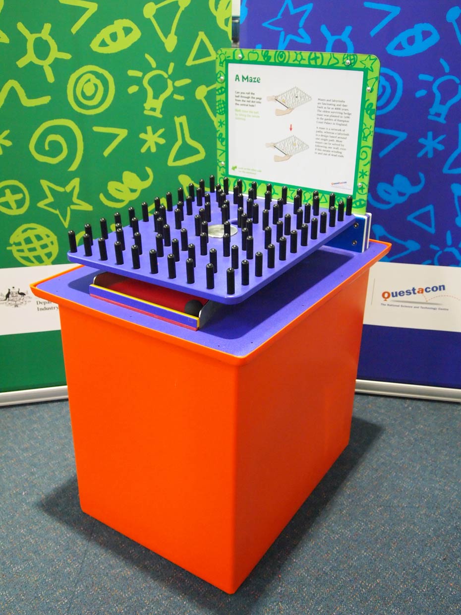 Tilt the tabletop to roll a ball around a maze of pegs and into a goal. An orange and purple exhibit table with a green and white information panel at the back, and a purple square plate on top of the table that has black pegs facing upwards.