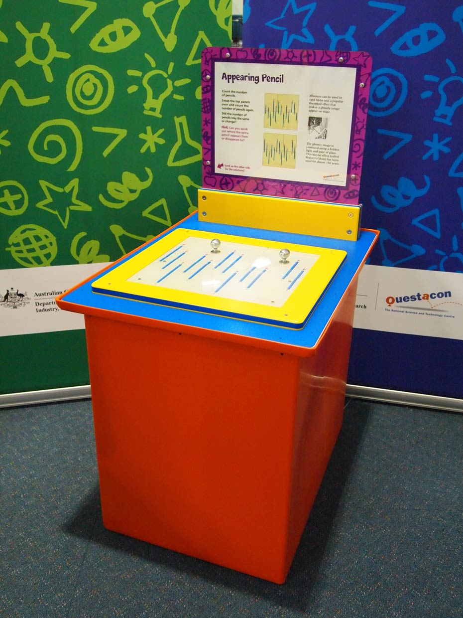 This visual illusion causes a pencil to appear, then reappear. A red and blue and yellow exhibit table with a purple and white information panel on top, sits in front of green and blue walls. On the table there are two brushed metal knobs with pictures of muliple blue pencils.