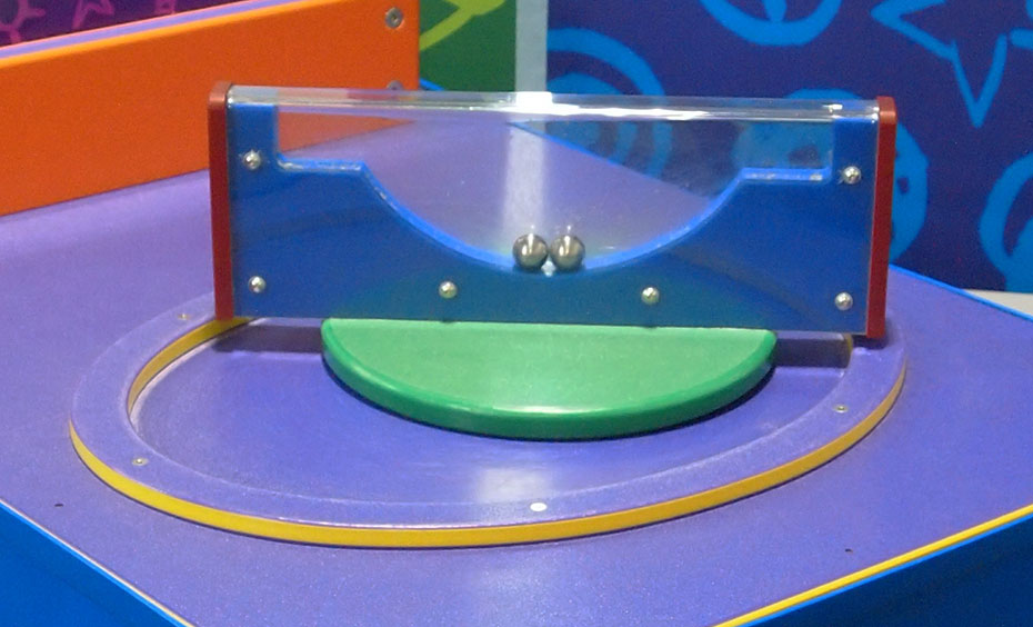 Try to perch two loose balls inside an enclosed container onto separate ledges, without touching the balls. A rectangular blue and clear perspex shape sitting on edge that has two silver balls inside.