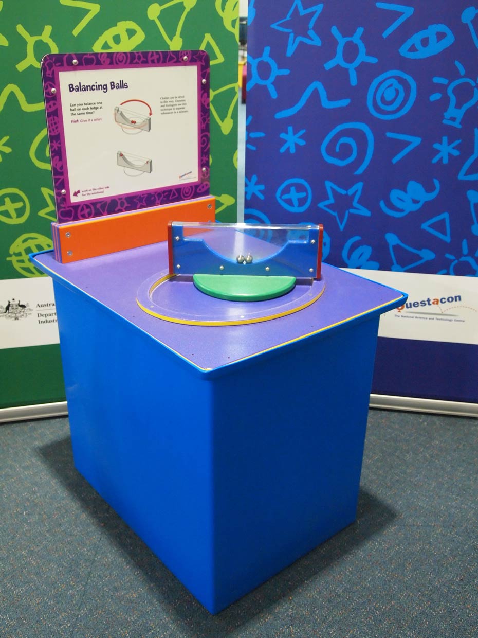 Puzzles can be solved using forces of physics. A blue and purple exhibit table with an orange, purple, and white information panel on top of it, sits in front of two green and blue walls. On the table is a rectangular blue and clear perspex shape sitting on edge that has two silver balls inside.