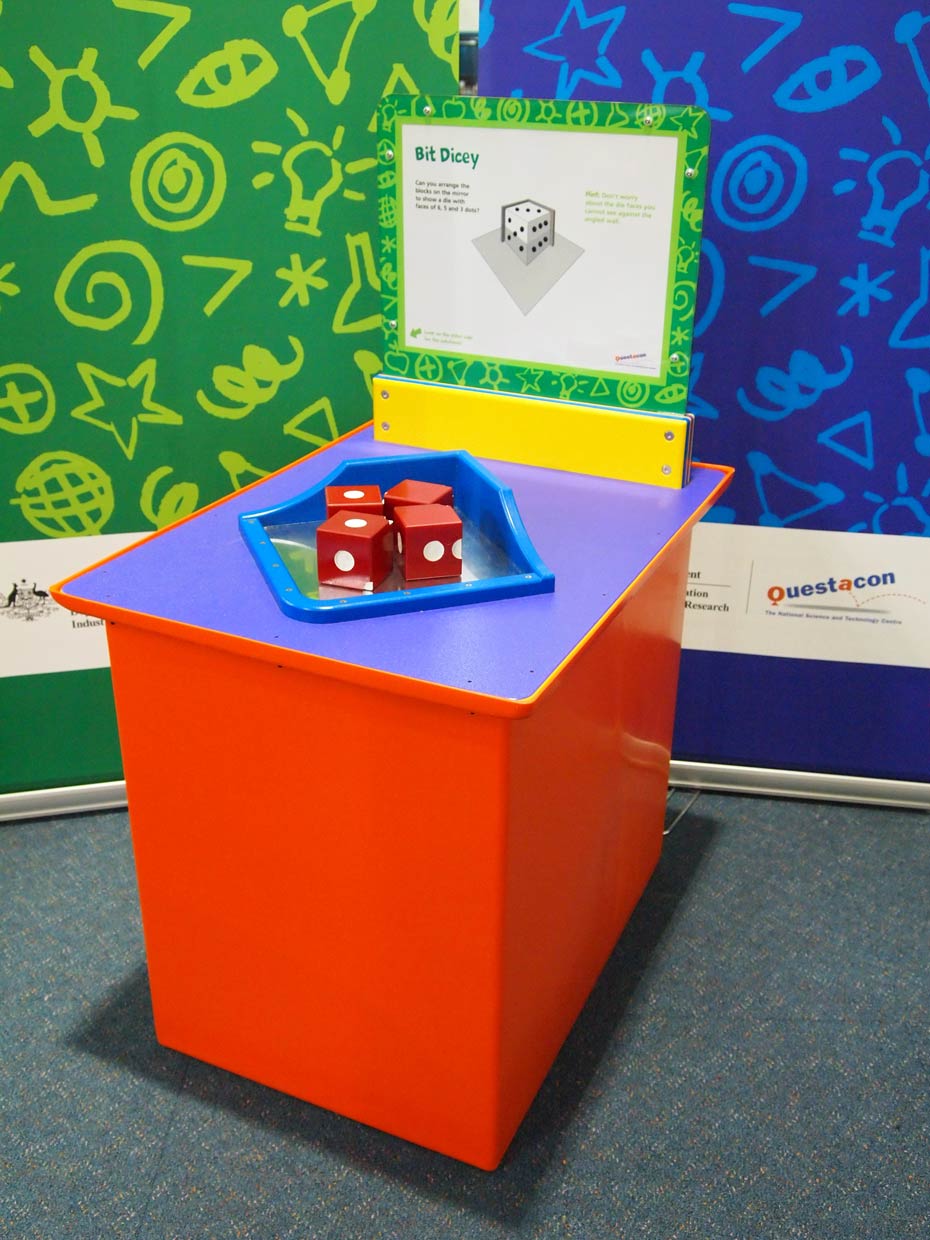 Reflections can be used to create a three dimensional image. A red and purple exhibit table, with a white, green and yellow information panel on top, sits in front of two green and blue walls. On the table sits four red and white die, sitting in a blue and reflective tray.