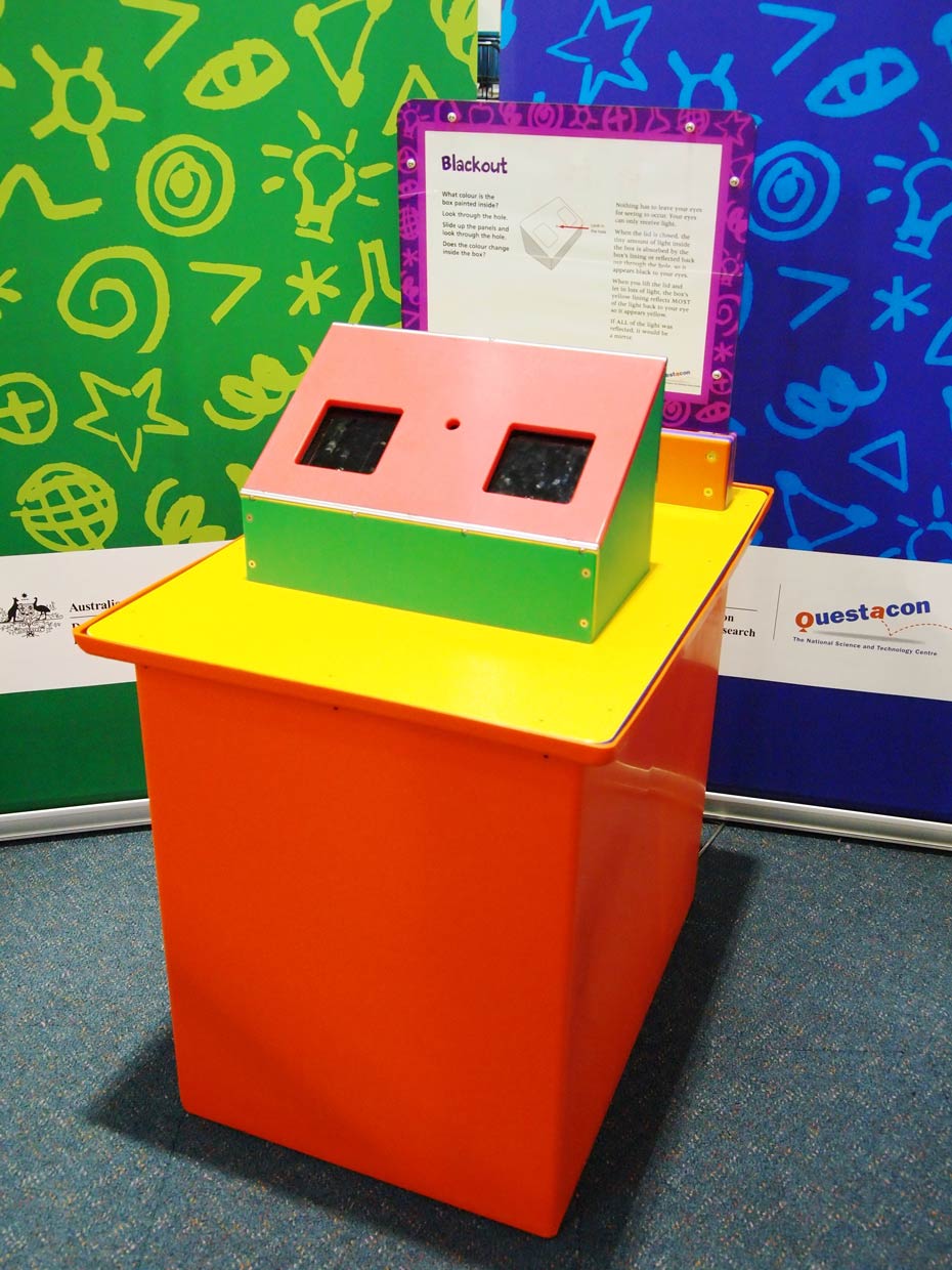 Humans can only see an object if light is being reflected from that object. A red and yellow exhibit table with an orange, white and purple information panel at the back, sits in front of two green and blue walls. On the table is a rectangular box with a sloped face and two square black areas.