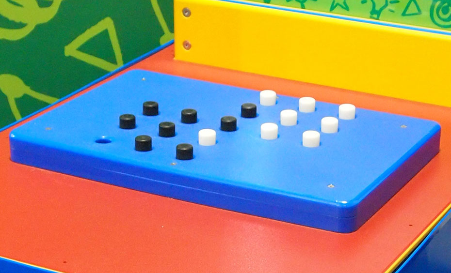 Black and white pegs need to be moved around a board in a minimum number of moves to create a new pattern. An exhibit table with a information panel, sits in front of two walls. On the table lays a rectangle shape with pegs sitting on top.