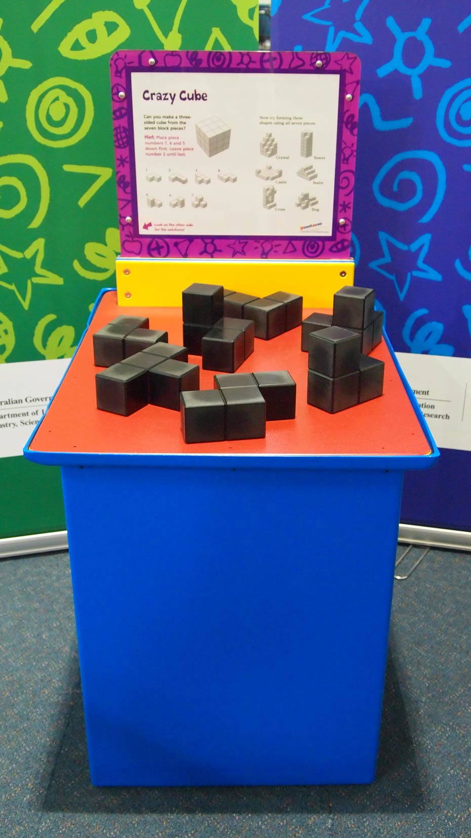 Make a cube from the seven block puzzle pieces. A blue and orange exhibit table with a yellow, white and purple information panel on top, sits in front of two green and blue walls. On top of the table are an array of small black cubes joined together in different shapes.