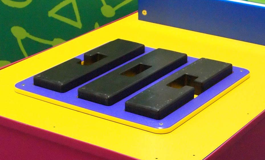 Three notched pieces of wood must be interlocked in sequence to create a three-dimensional cross. Three black shapes sit flat on a blue and yellow flat surface.