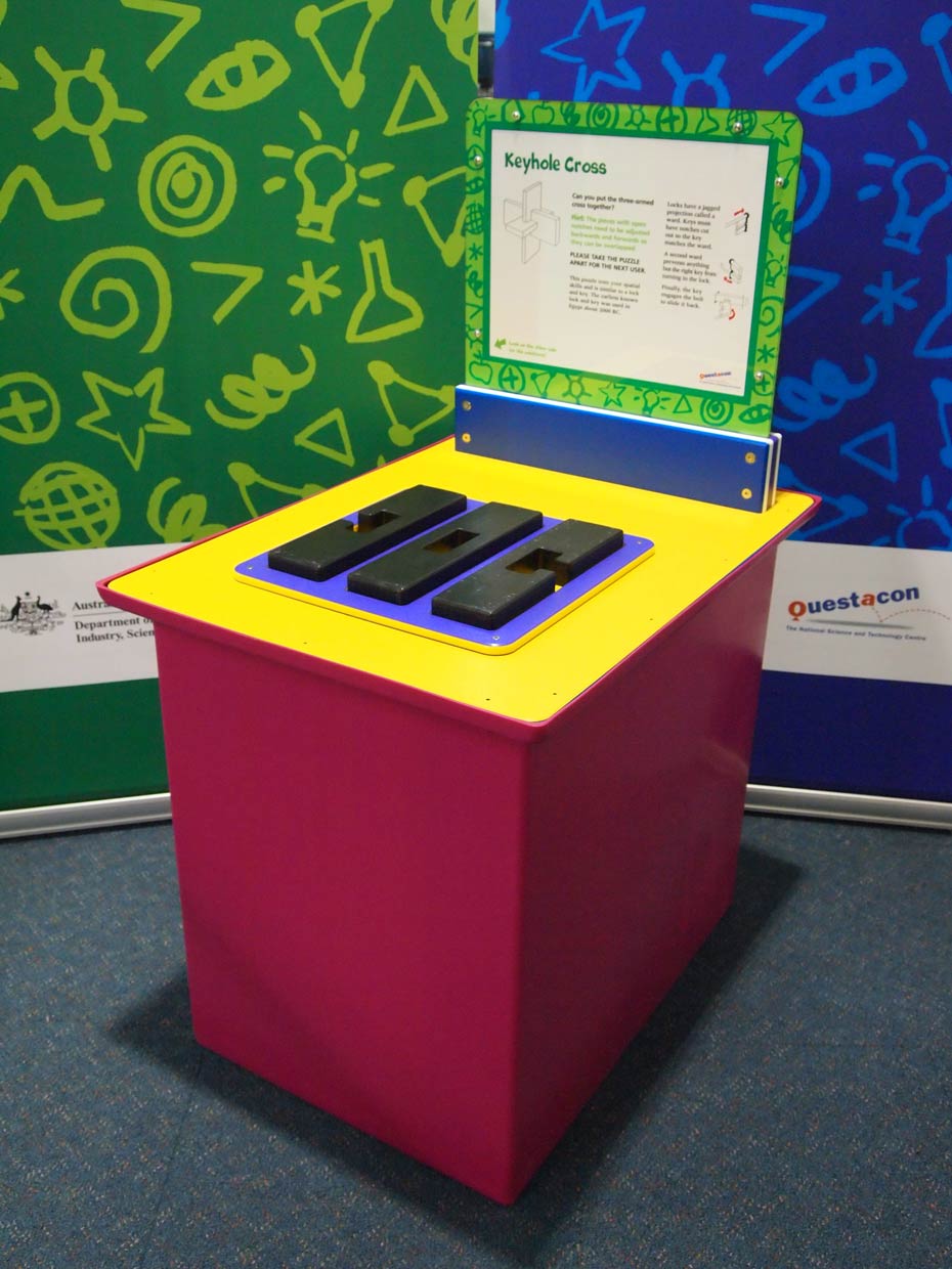 Structures can be built using 'lock and key' shapes. A red and yellow exhibit table with a blue, green and white information panel at the back, sit in front of two green and blue walls. On the table is a flat blue shape, with three black shapes on top.