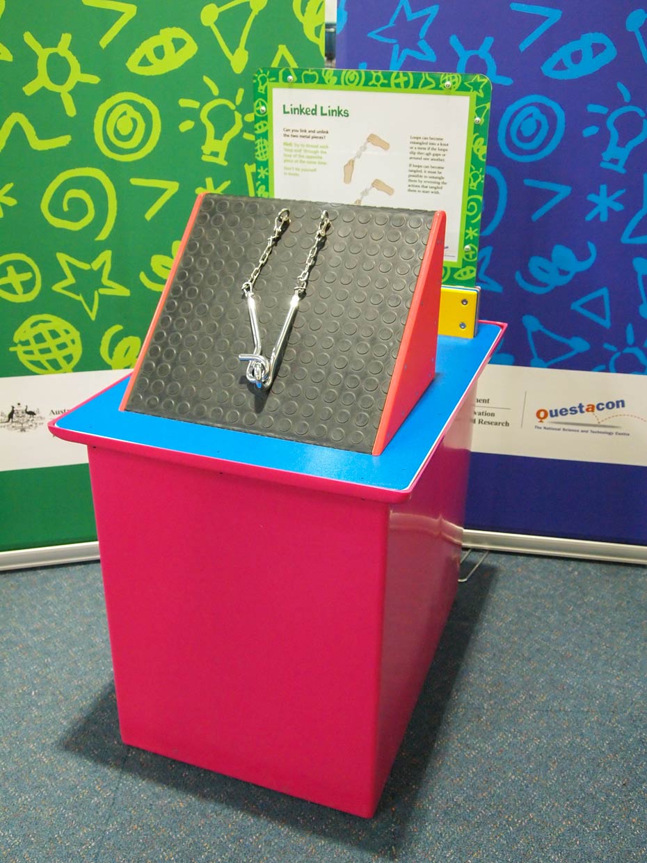 Topology puzzles explore patterns in space. A red and blue exhibit table, with a yellow, green and white informatiion panel on top, sits in front of two green and blue walls. On thed by chains to a black and red triangular prism. e table sits two stainless steel hoops attach