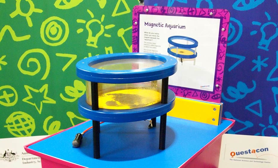 Slide a magnet across magnetite in a Perspex box and observe three dimensional magnetic fields. A red and blue exhibit table with a yellow, white and purple information panel on top, sits in front of two green and blue walls. On the table sits a blue, black and clear perspex cylinder shpaed aquarium.
