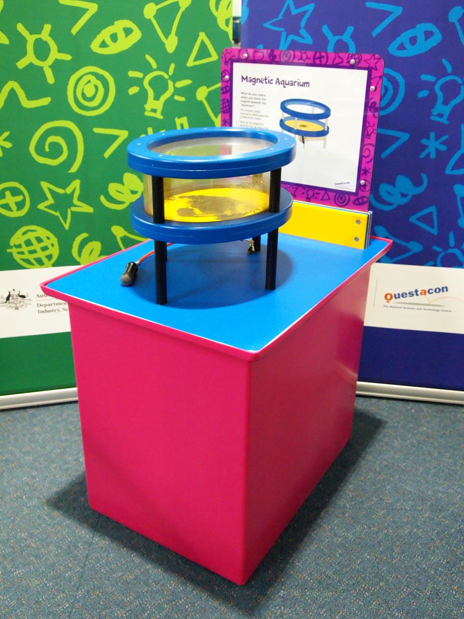 Magnetic fields can be seen when magnetic metals align against a magnet's poles. A red and blue exhibit table with a yellow, white and purple information panel on top, sits in front of two green and blue walls. On the table sits a blue, black and clear perspex cylinder shpaed aquarium.