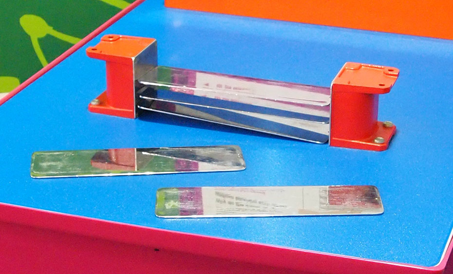 Stack the sheets of steel between the two strong magnets. Try taking the bottom sheet out and stacking it on top. A red and blue exhibit table with an orange, white and purple information panel on top, sits in front of a green and a blue wall. On the table top sits two orange end pieces that have flat chrome metal pieces suspended between them.