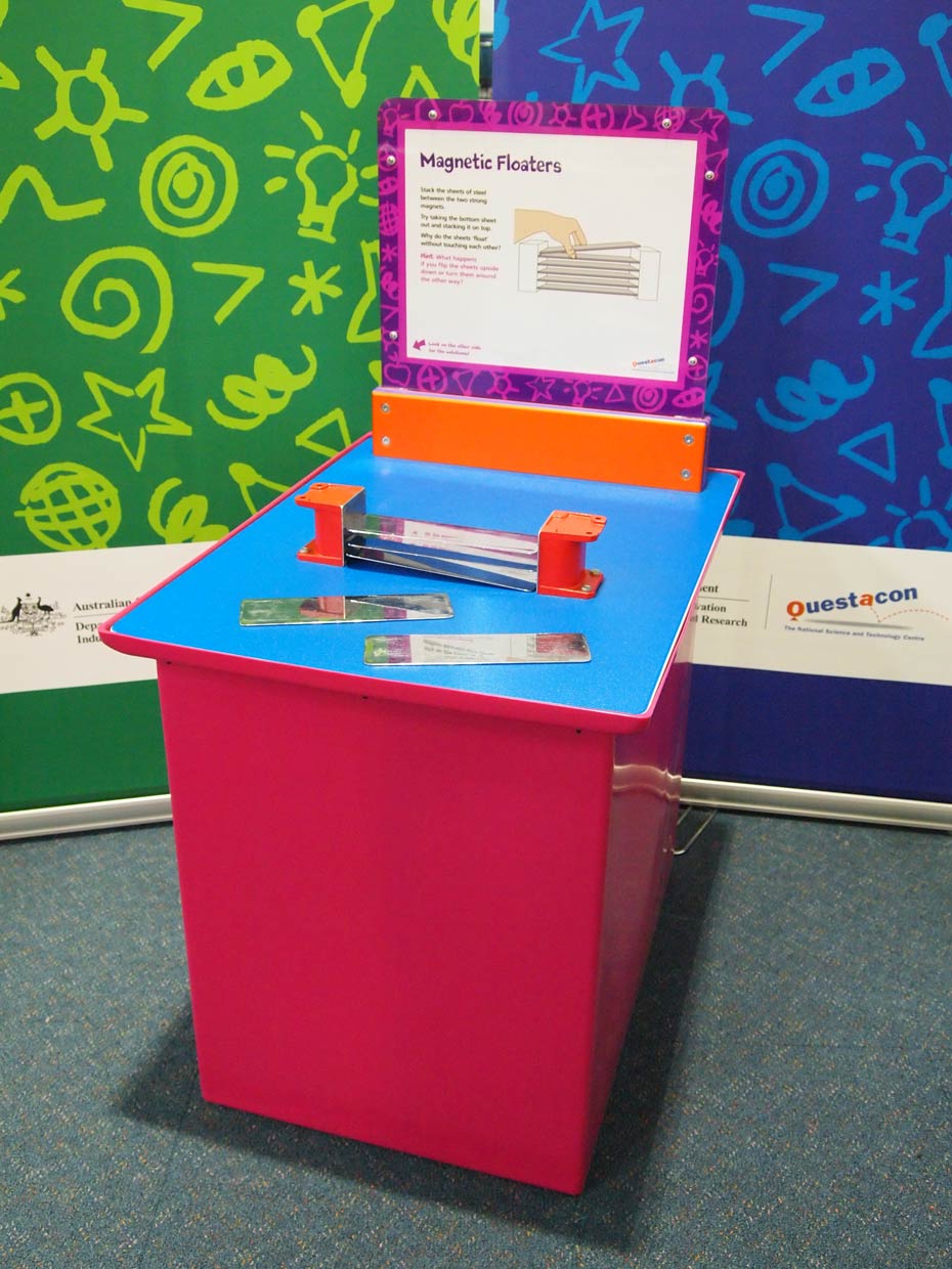 Metal plates (sandwiched between two powerful magnets) seem to float and repel each other when pushed down. A red and blue exhibit table with an orange, white and purple information panel on top, sits in front of a green and a blue wall. On the table top sits two orange end pieces that have flat chrome metal pieces suspended between them.