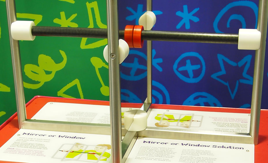 Watch your hand reflected in a mirror while it's sliding a plastic ring and notice whether the sliding hand reflection becomes confused with the hand hidden behind the mirror. Two stainless steel frames sit at right angles to one another, with some panels having mirrors and others air gaps. A white information panel is on the flat surface under the frames.