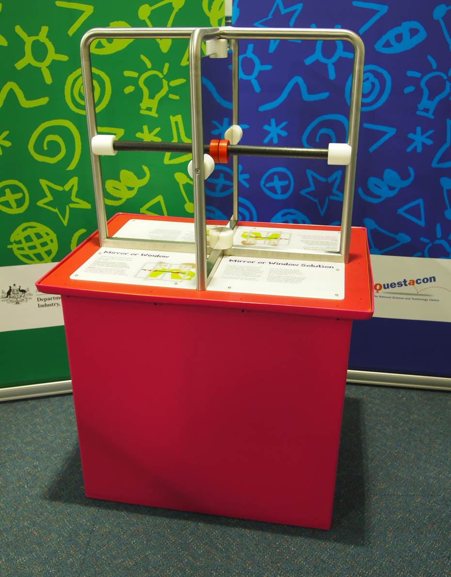 The human brain can be confused if visual signals do not match physical signals. A red exhiibit table with white infomation panels on the table top, sits in front of a green and a blue wall. On the table sits two steel frames at right angles to one another with mirrors and air gaps inbetween them.