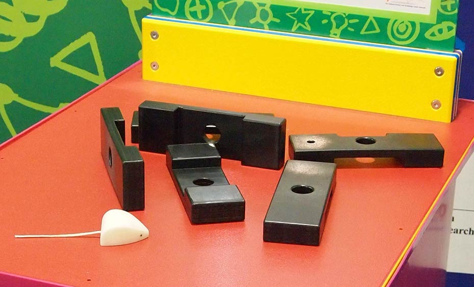 Slide various puzzle pieces together, so they interlock and form a three dimensional mouse mansion! A red and orange exhibit table, with a green, white and yellow information panel on top, sits in front of a green and a blue wall. On top of the table are five black blocks, and a toy white mouse.