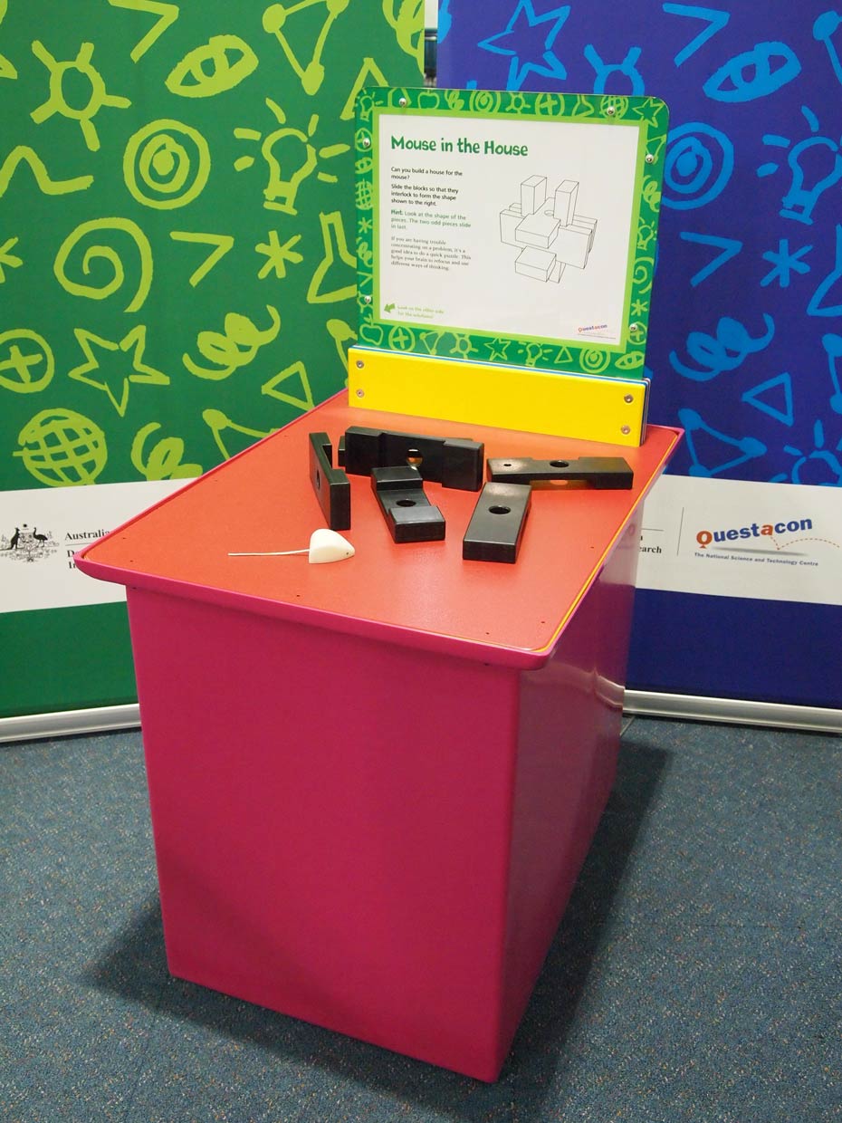 Structures can be built by interlocking pieces for strength. A red and orange exhibit table, with a green, white and yellow information panel on top, sits in front of a green and a blue wall. On top of the table are five black blocks, and a toy white mouse.