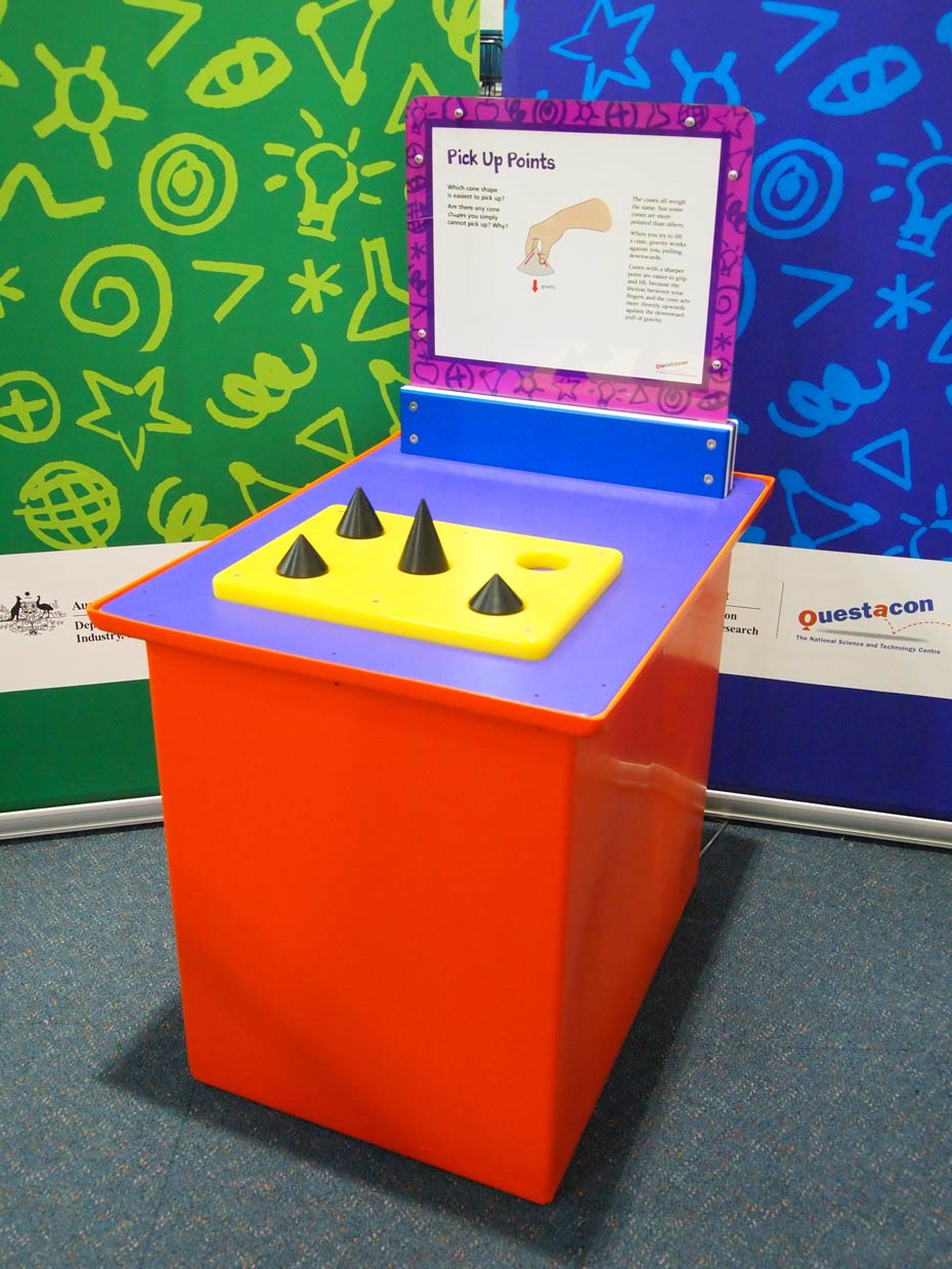 The force of friction allows items to be held and moved. A red and purple exhibit table, with a blue, white and purple information panel on top, sits in front of a green and a blue wall. On the table is a yellow flat board, with four black cones pointing upwards.