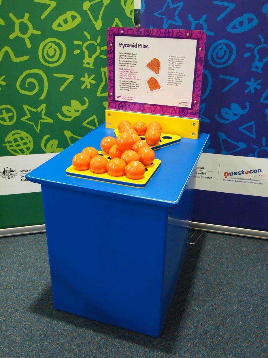 Build a pyramid with a triangular or rectangular base, using the same siz puzzle pieces of joined balls. A blue exhibit table, with a yellow, white and purple information panel on top, sits in front of a green and a blue wall. On the table are a triangle shaped and a rectangle shaped yellow flat base, on which sits piles of orange balls.