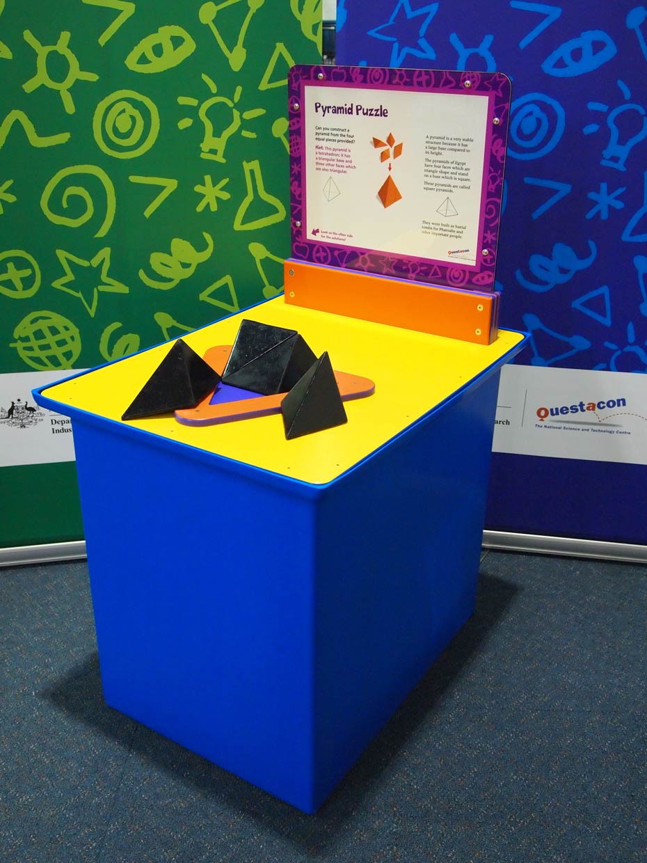 Try to build a large pyramid using the pyramid-shaped blocks. A blue and yellow exhibit table, with an orange, white and purple information panel on top, sits in front of a green wall and a blue wall. On top of the table sits a flat orange triangle base and four black triangular pieces.