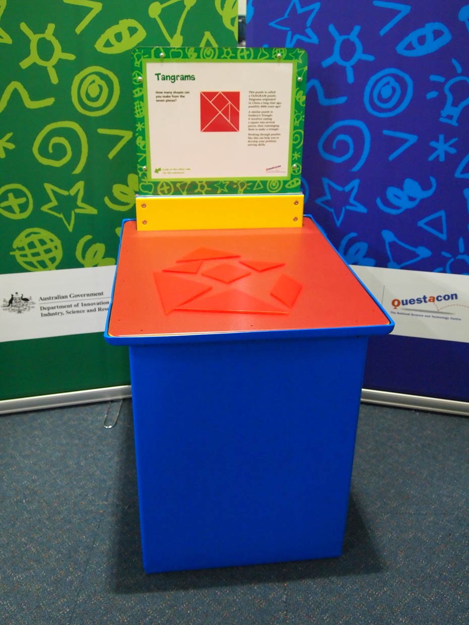 Try your hand at this ancient puzzle and mae a variety of shapes from the same seven puzzles pieces. A blue and orange exhibit table, with a yellow, green and white information panel on top, and a green and a blue wall behind. On top of the table are a range of differnt flat orange shapes.