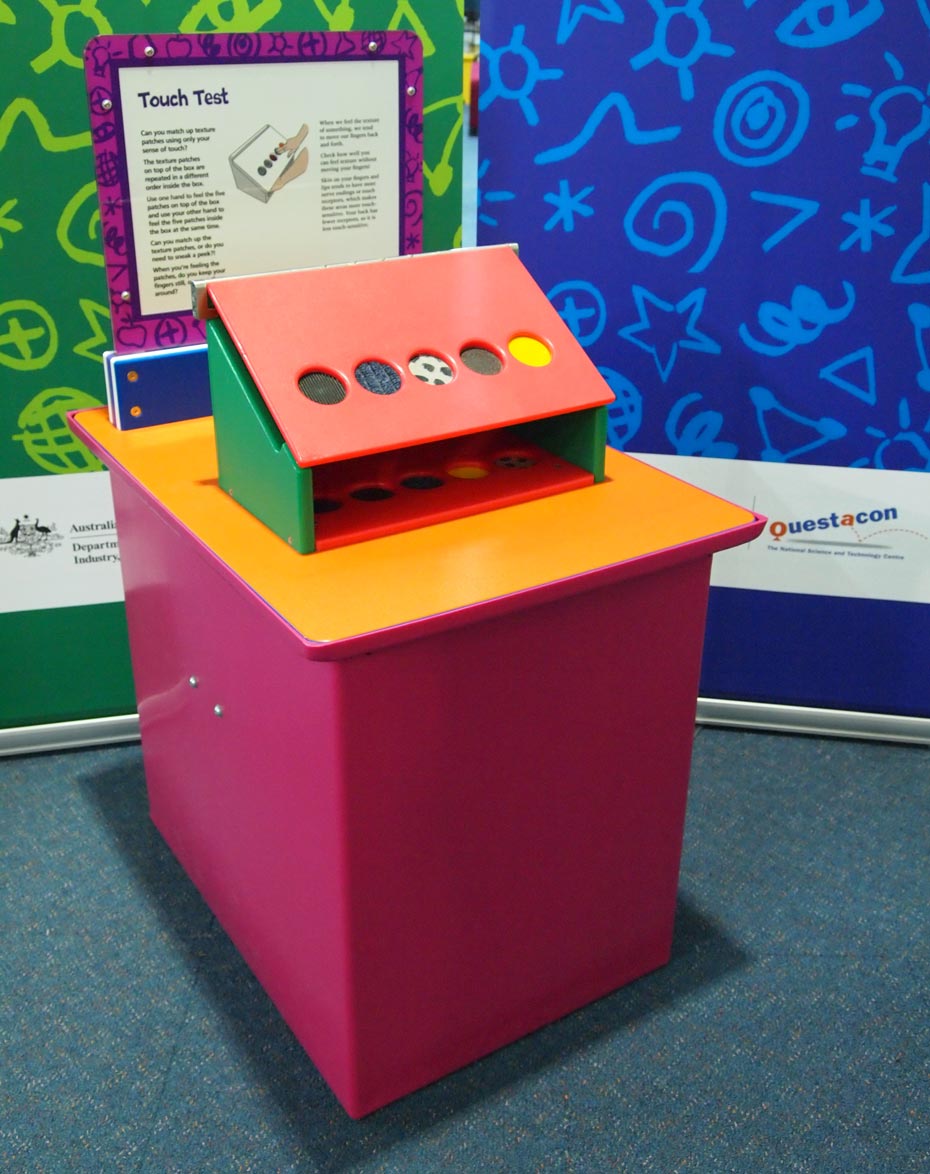 Touch different textured patches and match them to hidden texture patches. A pink and yellow exhibit table, with a blue, purple and white information panel on top, and a green and a blue wall sitting behind. On the table sits a box with a sloped top face that has five circular cut outs, that each have a different fabric.