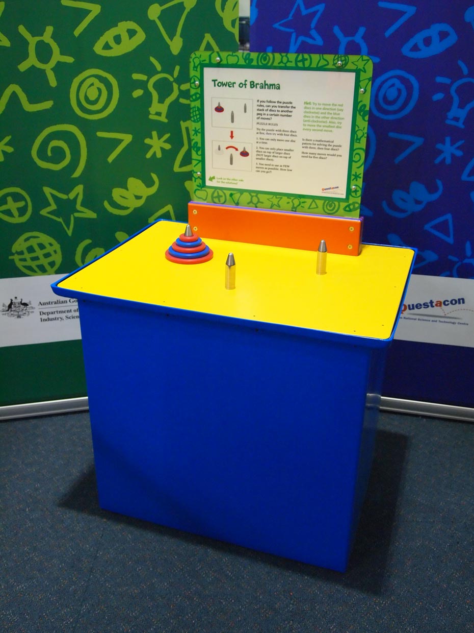 Is there a mathematical pattern for solving the puzzle with three, then four discs? A blue and yellow exhibit table, with a green, white and orange information panel on top, sitting in front of a green and a blue wall. On the yellow tabel top are three chrome pegs, of which one has five red and blue discs that are threaded onto the peg, and arranged from largest to smallest.