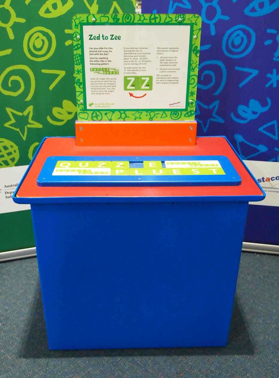 Movement of tiles in a puzzle represent processes of translation and rotation. A blue and red exhibit table, with a green, white and orange information panel on top, sits in front of a green and a blue wall.