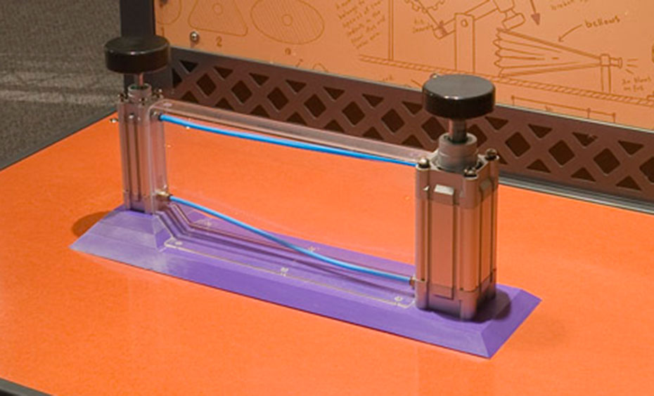 Push down the narrow piston and watch how the attached, wider piston moves, due to transferred pressure and force over an area. A black and orange exhibit table, with matching information panel at the back. On the table sits two silver pistons with black handles and a purple base between them.