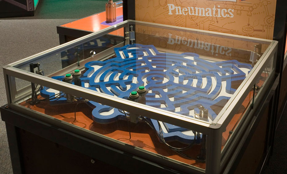 A team of people use pneumatic pistons and problem solving skills to roll a ball through the tilting maze. A large blue and white flat maze that is contained within a blass and steel display case, sits in front of a brown and white vertical information panel.