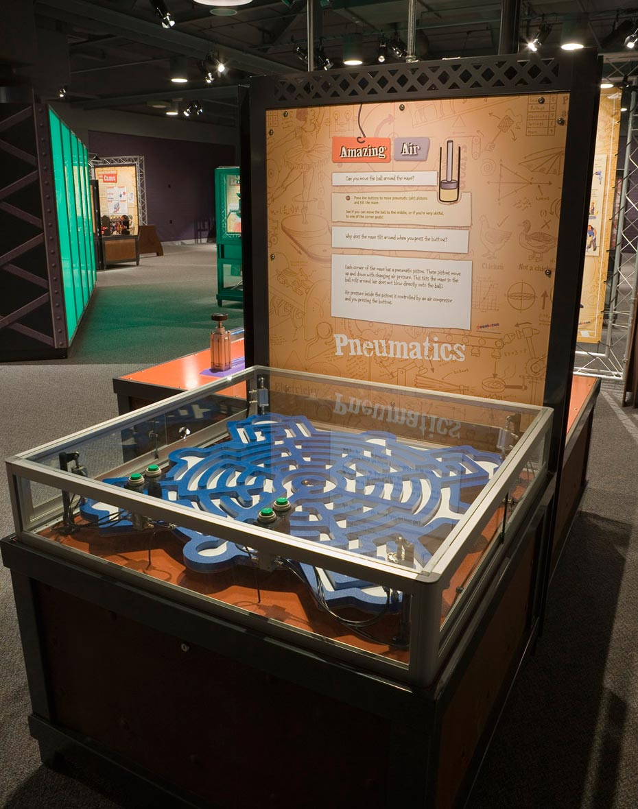 Pneumatic pistons use air to lift and lower heavy things. A large blue and white flat maze that is contained within a blass and steel display case, sits in front of a brown and white vertical information panel.