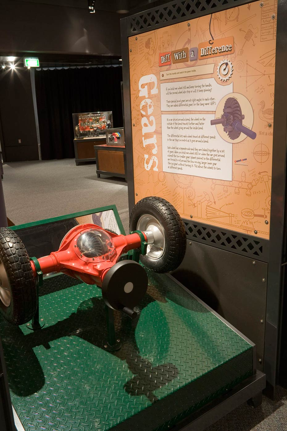 Simple machines work together to steer a car. A black and green display base that has a red, car differential with a see-through window to show the gears. To the right is a brown and white information panel.