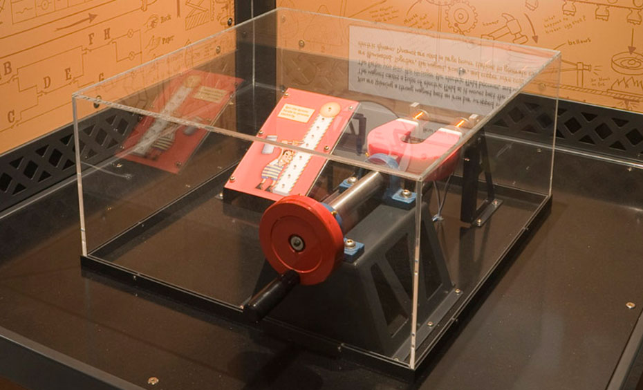 Spin a magnet past an iron bar wrapped in copper wire to generate an electric field and illuminate lights on a meter. A red mechanical dynamo device enclosed in clear pespex sits on a black bench, that is surrounded on two sides by brown and white information panels.