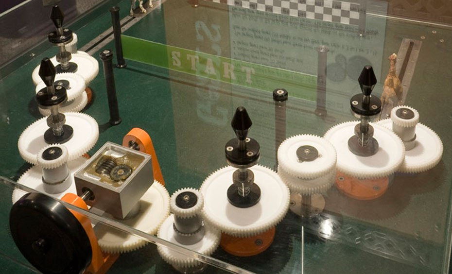 Combine small, medium and large gears and turn a handle to race a toy camel and a toy horse along a track. A black exhibit table with a clear perspex box over it, enclosing a series of white gears all connected together.